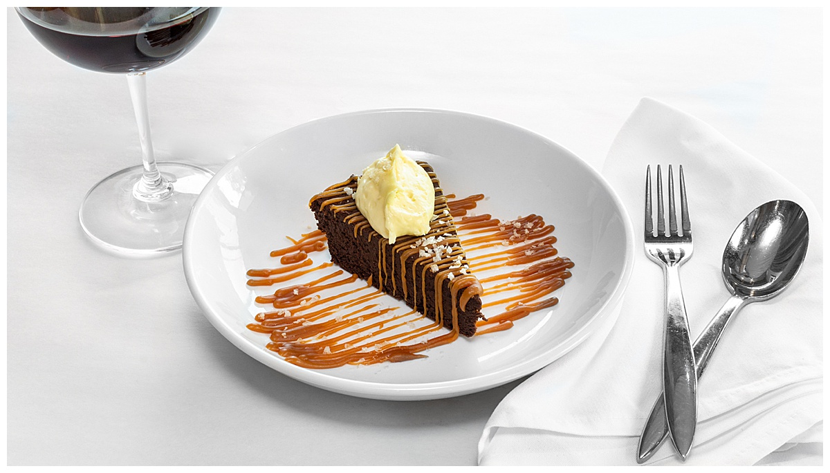 A dessert sits on a white plate with a white tablecloth, white napkin, wine, and silverware around it.