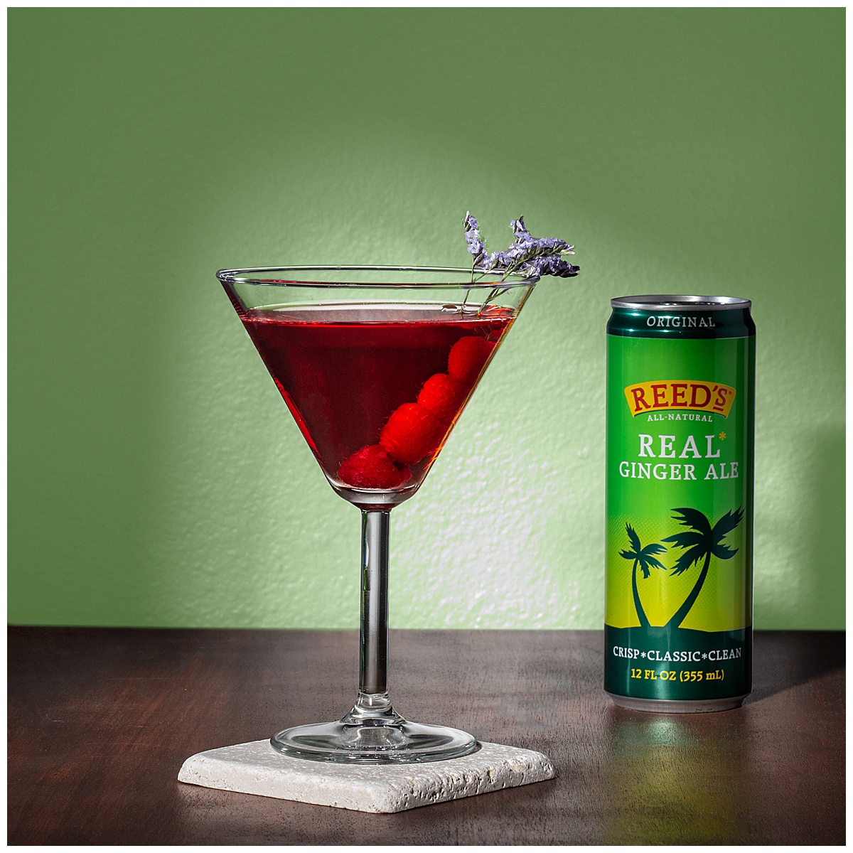A red martini with a purple flower sits on a stone coaster with an unopened can of Reed's Real Ginger Ale. They're sitting on dark wood with a green background.
