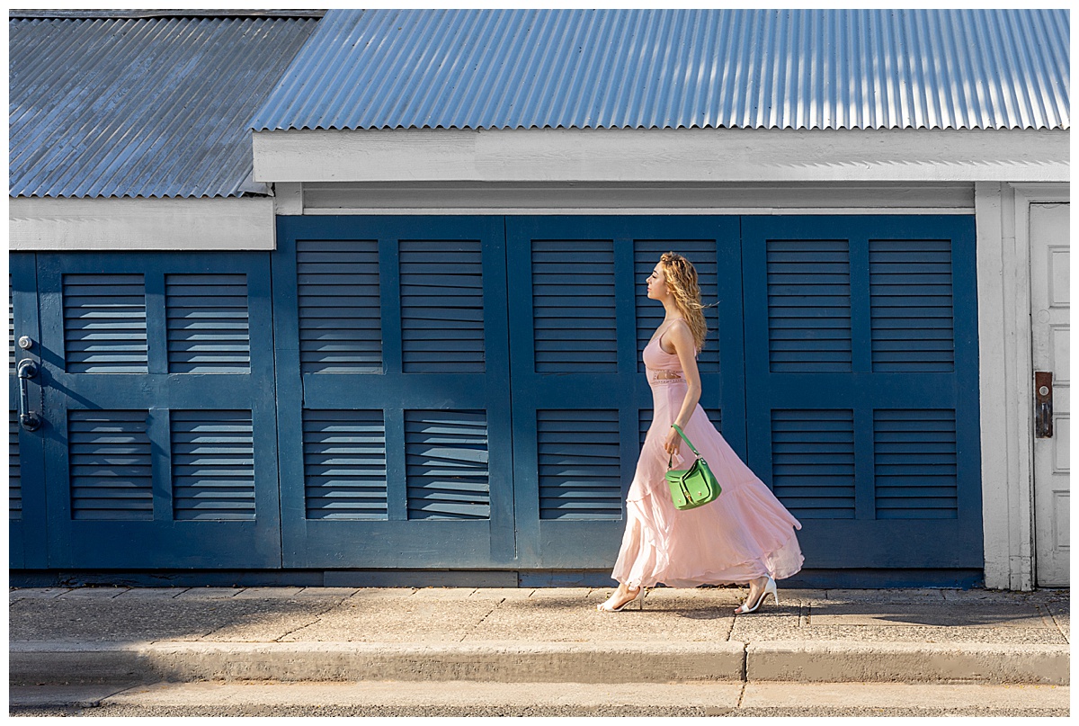 Woman with curly blonde hair wearing a pink dress walks in front of a blue wall with shutters holding a green purse posing in a fashion photoshoot.