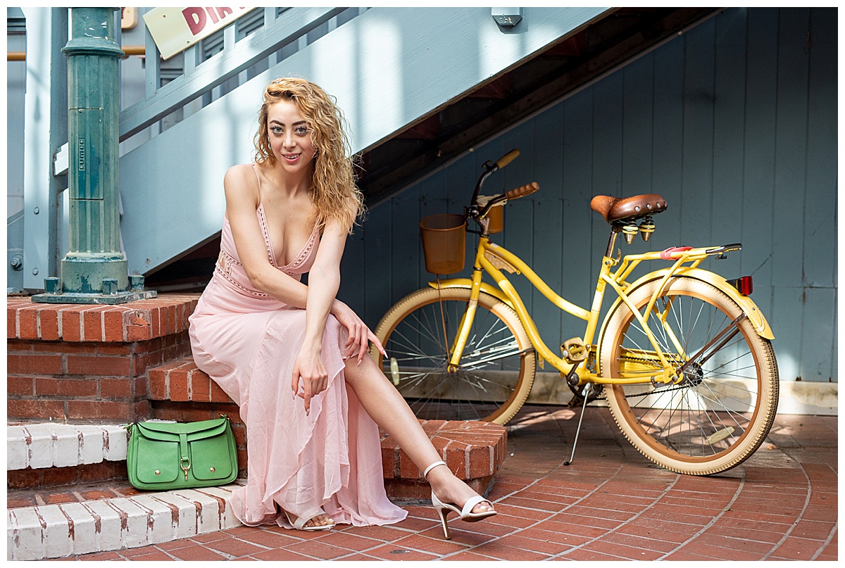 Woman with curly blonde hair wearing a pink dress sits on a brick step in front of a yellow bicycle with a green purse.