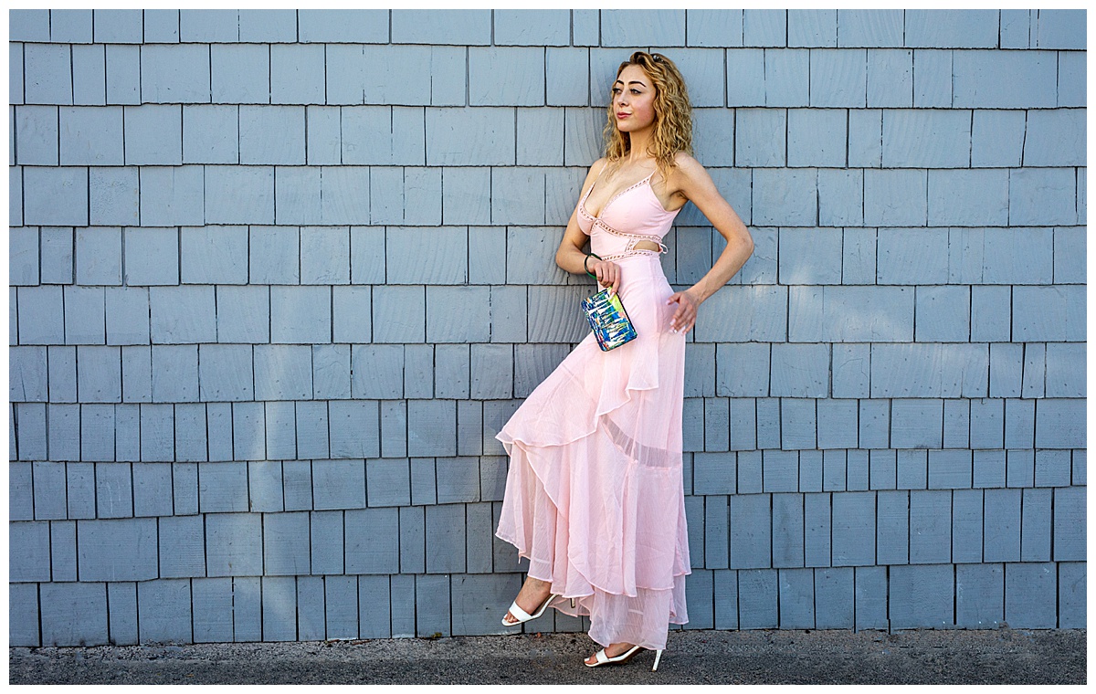 Woman with curly blonde hair wearing a pink dress leans against a blue shingled wall holding a blue purse with boats on it.