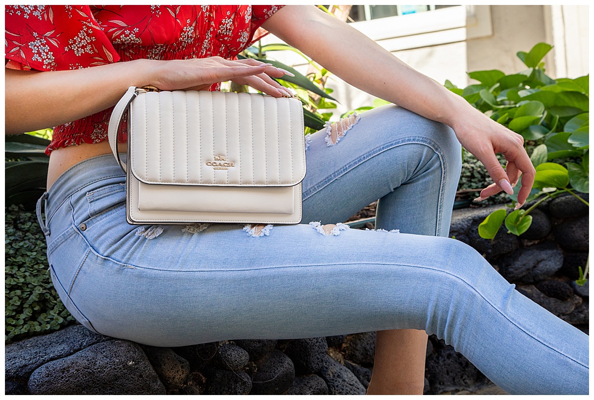 A close up of a woman wearing a red shirt and blue jeans holding a white Coach purse posing in a fashion photoshoot.