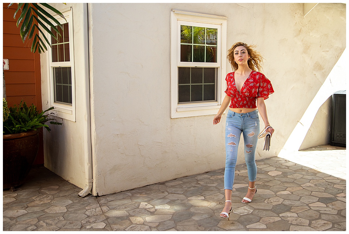 Woman with curly blonde hair wearing a red shirt struts towards the camera with a white Coach purse posing in a fashion photoshoot.