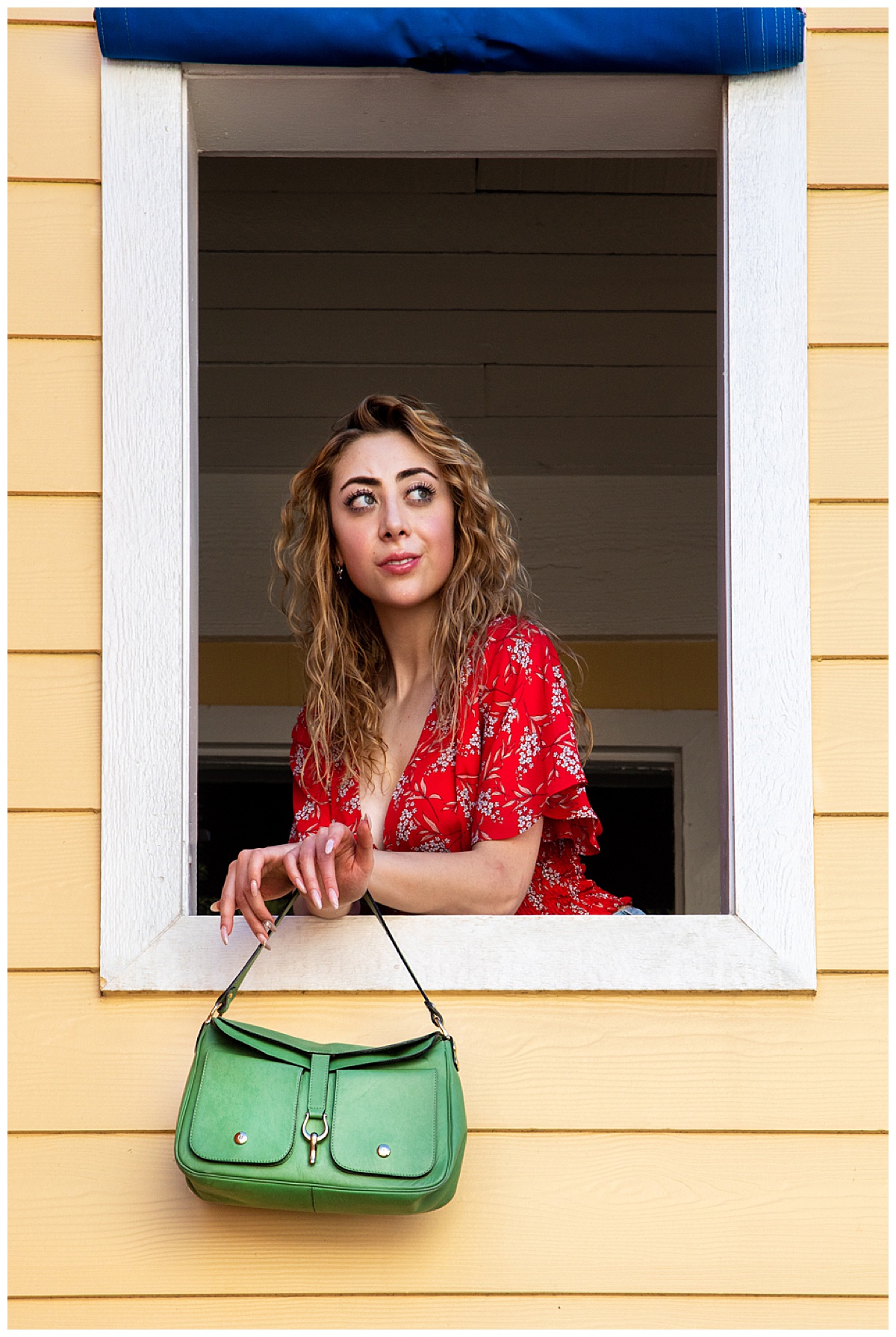 Woman with curly blonde hair wearing a red shirt leans out of a yellow window holding a green purse posing in a fashion photoshoot.