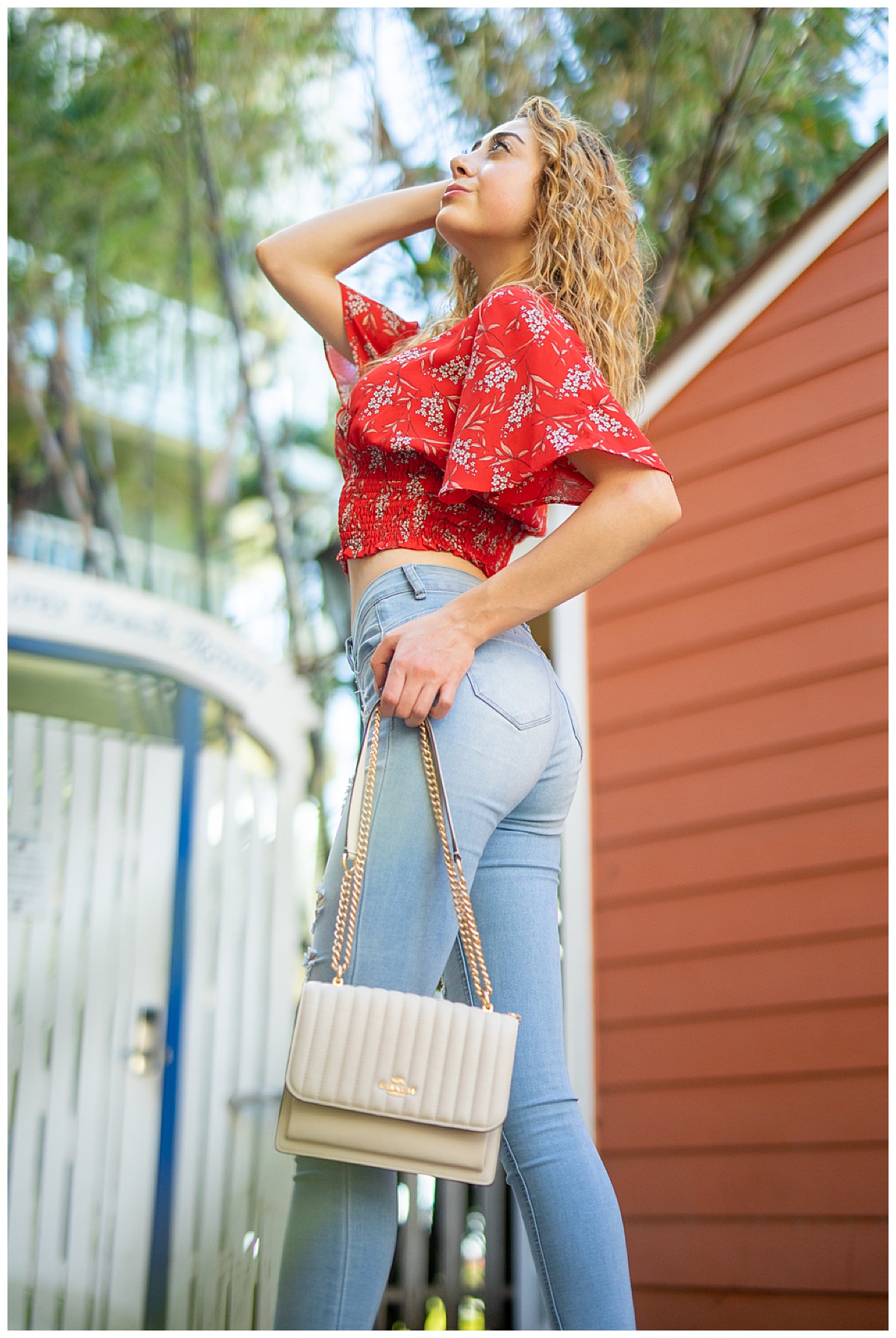 Woman with curly blonde hair wearing a red shirt holds a white Coach purse posing in a fashion photoshoot.