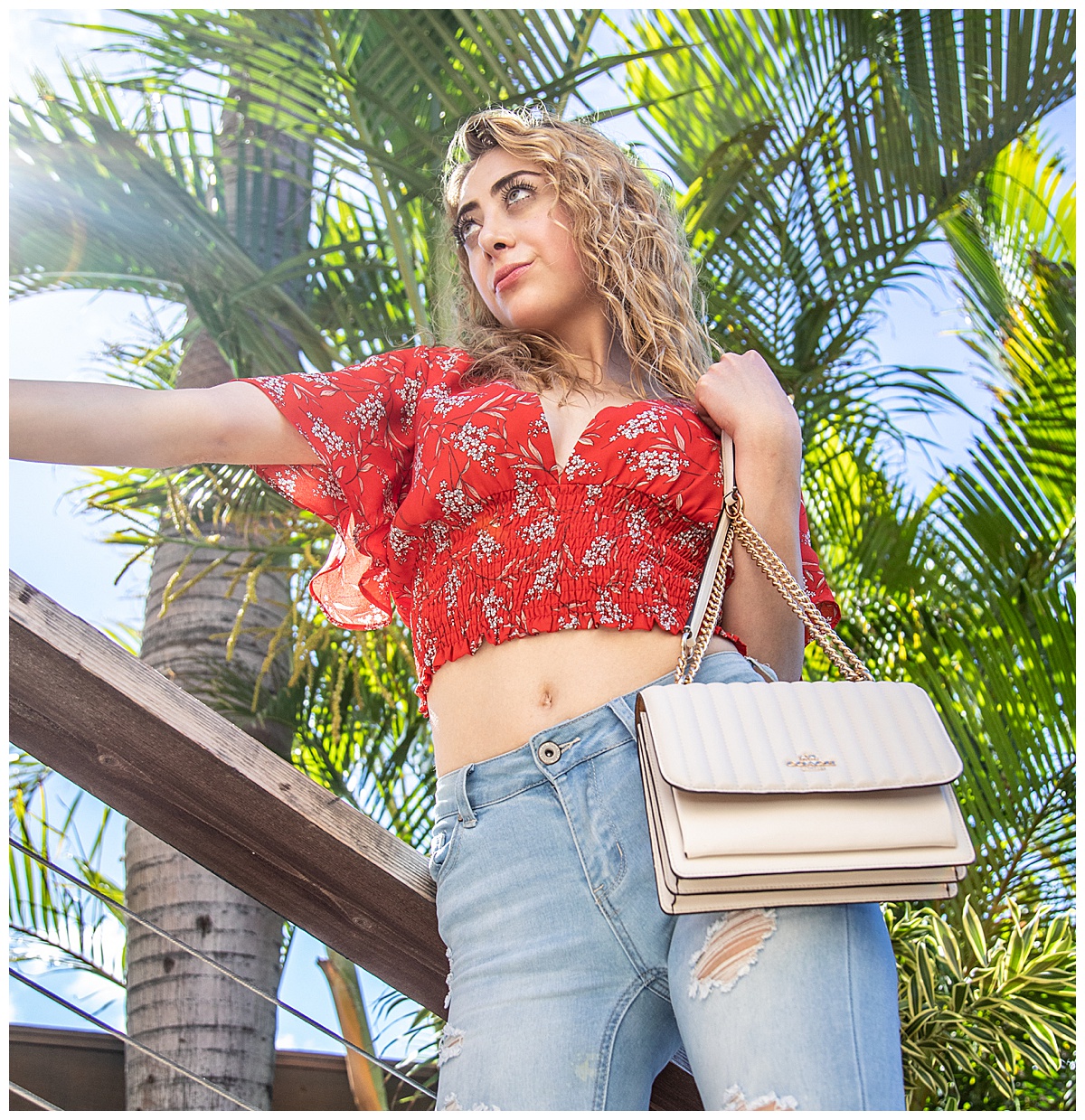 Woman with curly blonde hair wearing a red shirt holding a white Coach purse posing in a fashion photoshoot.
