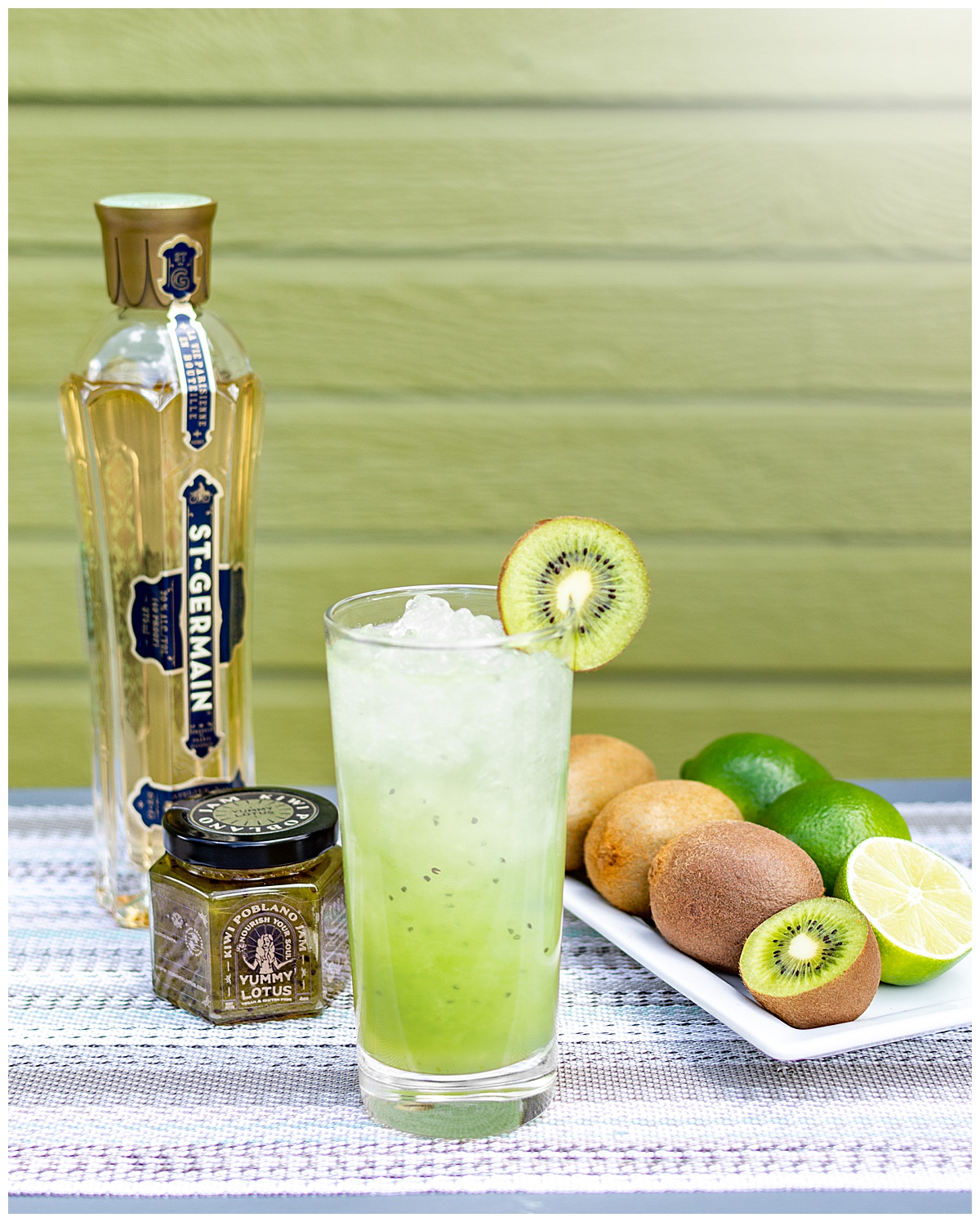 A photo showcasing a Yummy Lotus jam & cocktail pairing. A tall green drink with ice and a kiwi, the Kiwi Poblano jam, a bottle of St Germain liqueur, and a plate of kiwis and limes are on a placemat in front of a green wall.