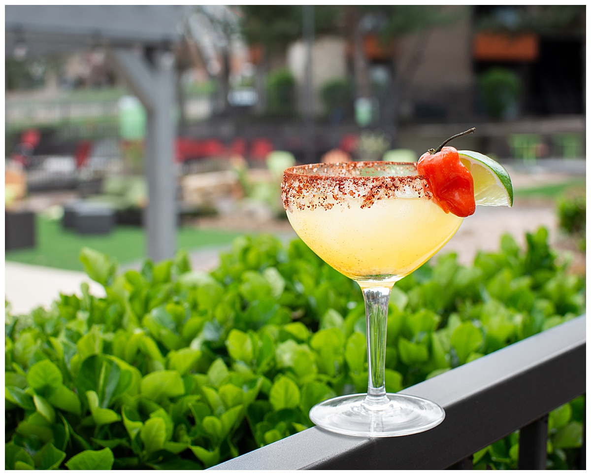 A photo showcasing a Yummy Lotus cocktail pairing. A yellow, spicy margarita with a salted spicy rim, a pepper, and a lime, sits on a railing in front of a bush. A large, fun seating area is out of focus in the background.