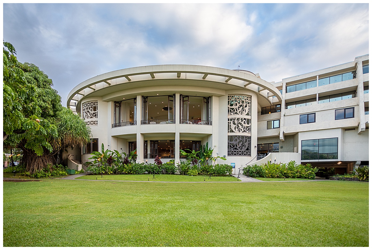 The outside of the resort. The building is white and circular with lots of windows. There is a big green lawn in front with lots of plants.