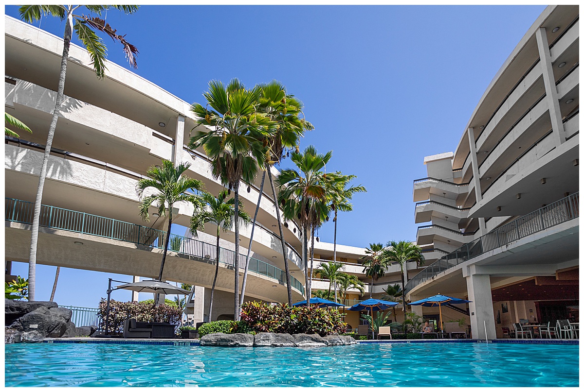 View of the Outrigger Kona Resort & Spa from the adults only pool. There are lots of palm trees and blue umbrellas.