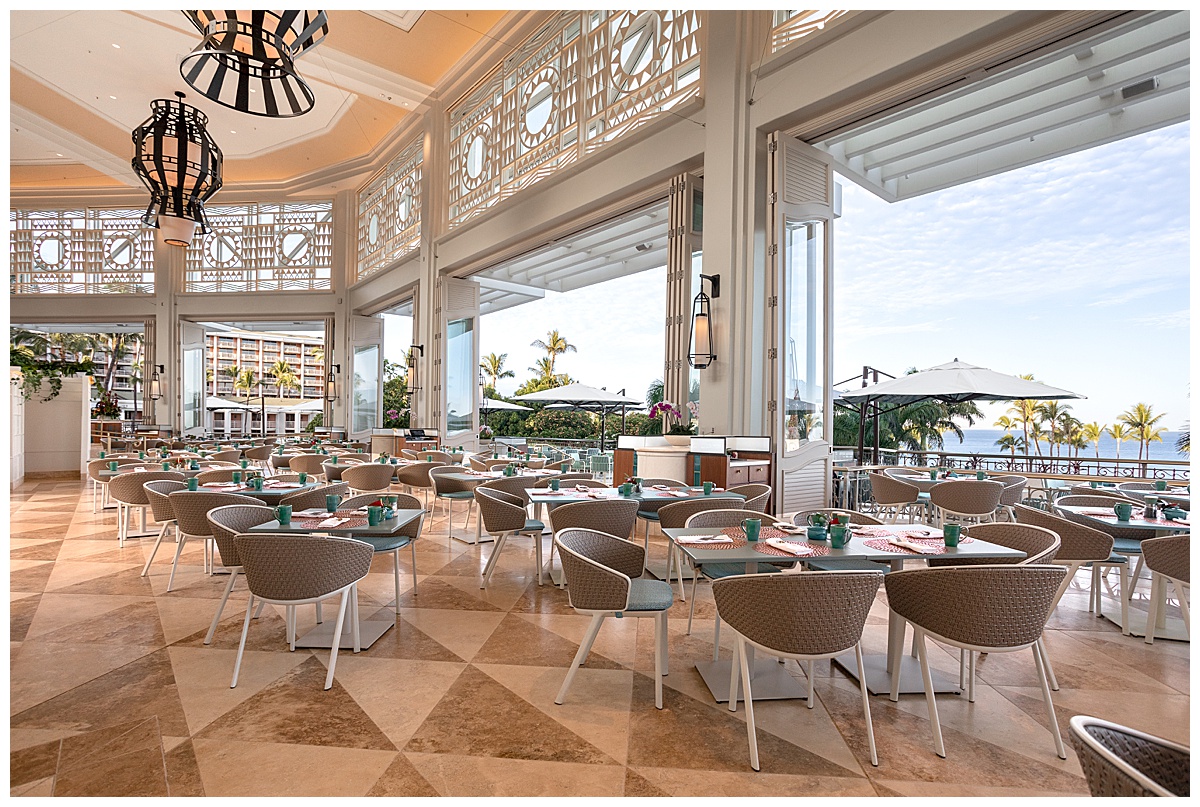 The seating area of the restaurant. The white walls are scarce as the restaurant is very open air. The tables are light teal squares and rectangles, and the chairs are rounded with brown wicker backs. The floor is a brown marble triangle tile.